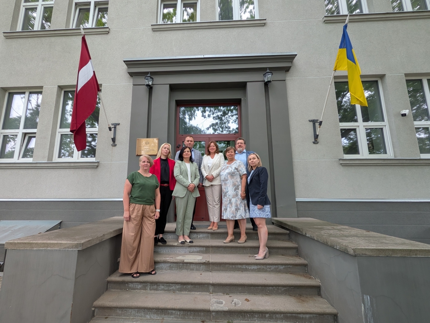 Latvijas un Ukrainas kolēģi stāv uz kāpnēm pie Valsts augu aizsardzības dienesta ieejas, fonā Latvijas un Ukrainas karogi