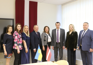 8 cilvēku grupas bilde, priekšplānā uz galda Ukrainas un Latvijas karodziņš, fonā liels Latvijas karogs