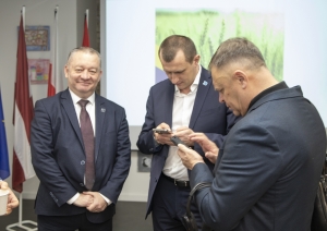 Trīs uzvalkos ģērbri vīrieši, diviem rokas telefonu, kaut ko raksta