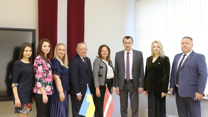 8 cilvēku grupas bilde, priekšplānā uz galda Ukrainas un Latvijas karodziņš, fonā liels Latvijas karogs