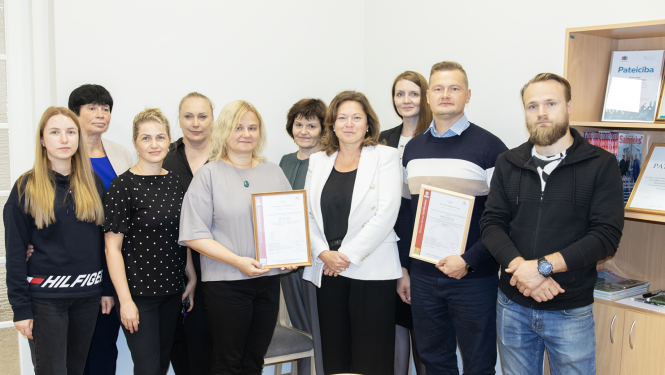 9 cilvēki, kopbilde, divi tur rokās ierāmētus sertifikātus