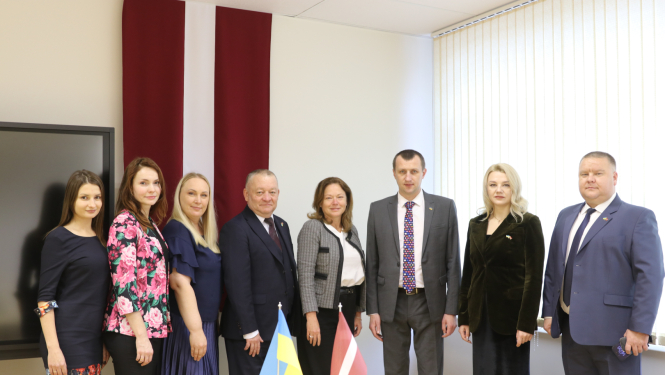 8 cilvēku grupas bilde, priekšplānā uz galda Ukrainas un Latvijas karodziņš, fonā liels Latvijas karogs