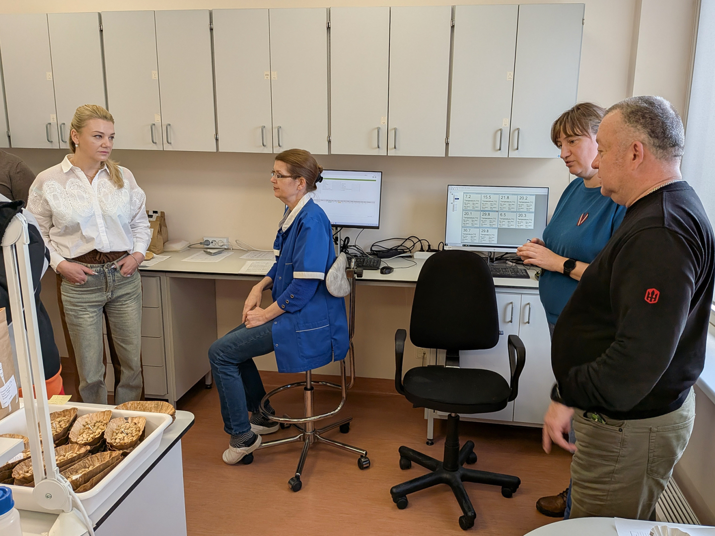 Sēklu dīgtspējas laboratorijas telpas, eksperte sēž, divi cilvēki stāv, priekšplānā paraugi un aprīkojums