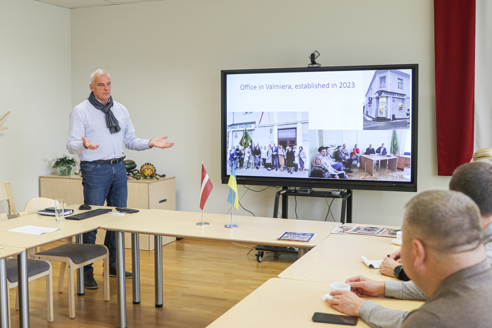 Ringolds Arnītis zālē stāsta prezentāciju, fonā ekrānsm, priekšplānā pie galda sēž cilvēki