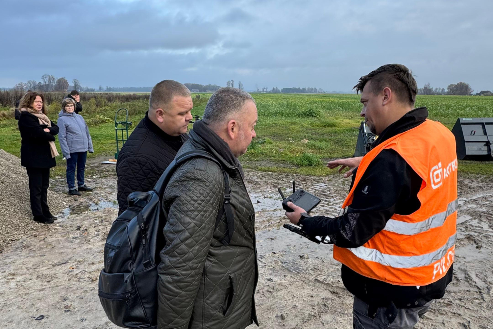 Priekšplānā 3 cilvēki - lauksaimniecības dronu pilots demonstrē Ukrainas kolēģiem dronu vadības pulti - nelielu monitoriņu