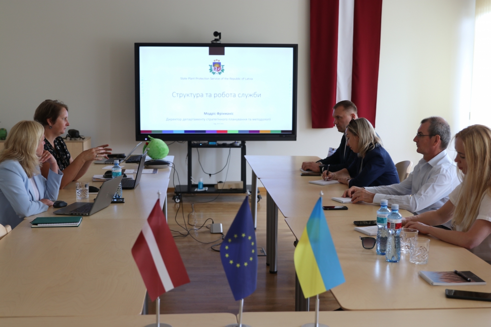 Cilvēki zālē pie galdiem, skatās prezentāciju, priekšplānā LV, ES un UA karogi 