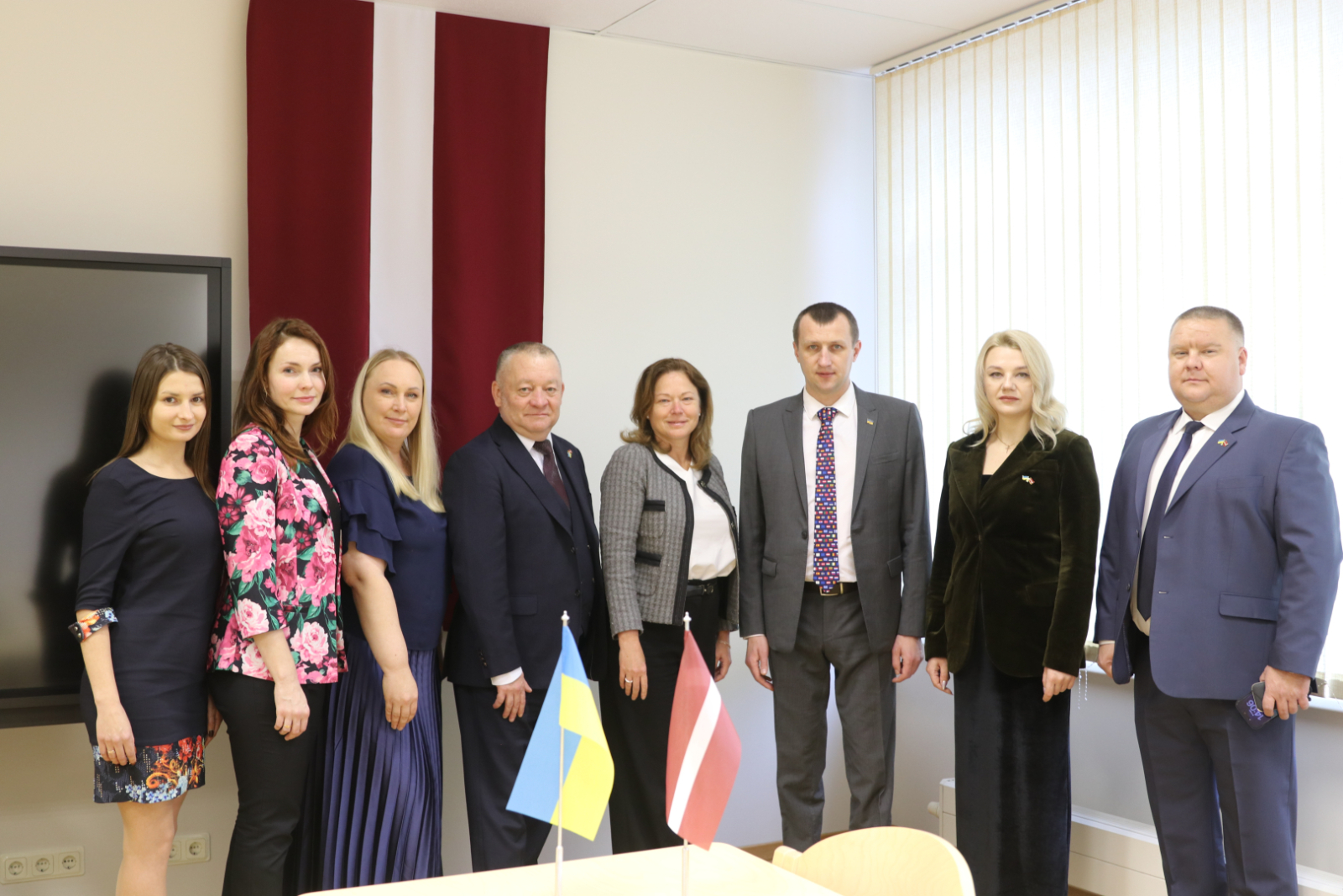 8 cilvēku grupas bilde, priekšplānā uz galda Ukrainas un Latvijas karodziņš, fonā liels Latvijas karogs