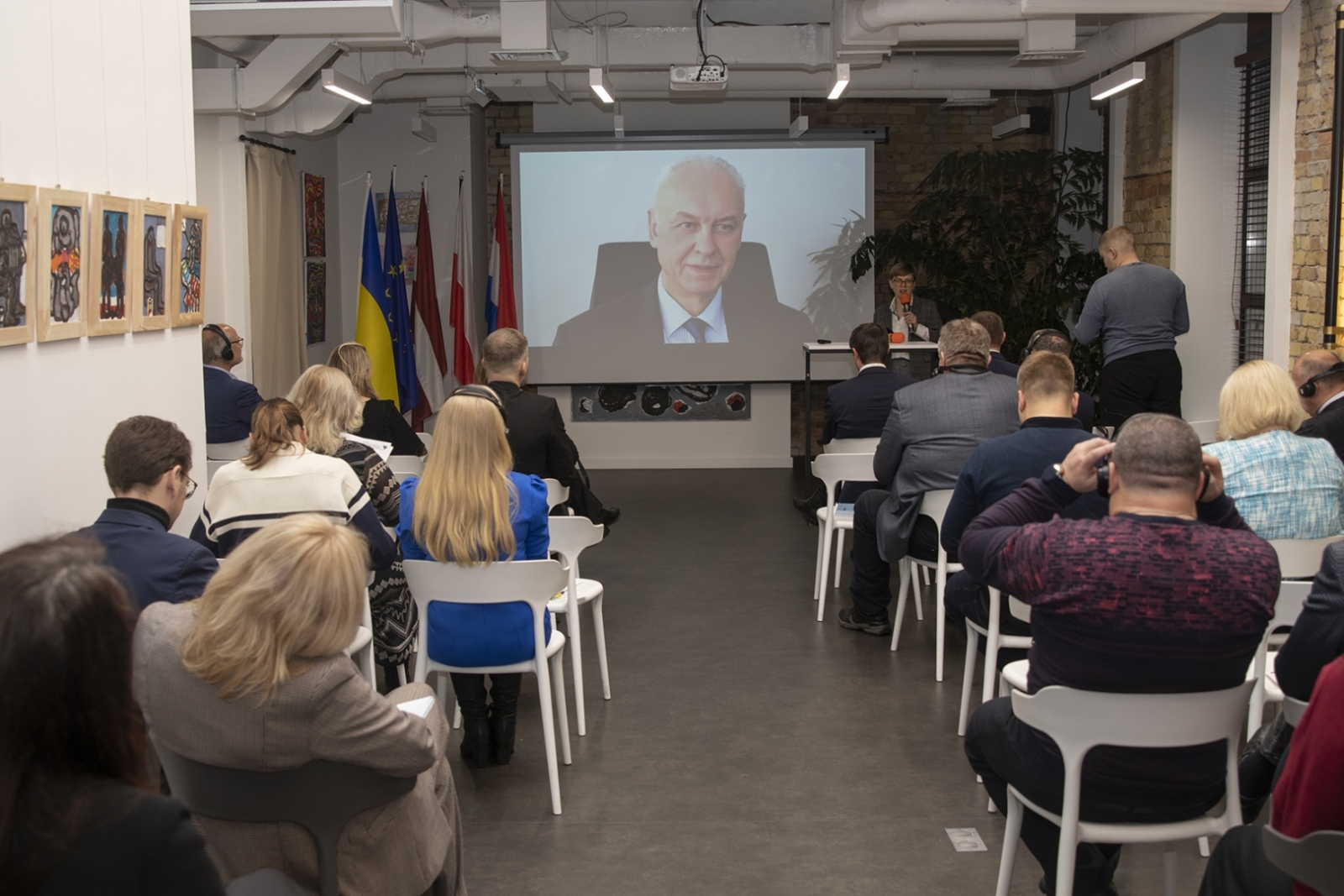 Skats no telpas dziļumu, cilvēki sēž zālē, priekšā ekrāns, kurā redzams vīrietis, kas saka runu